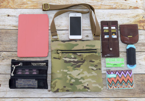MultiCam® Crossbody Bag shown against wood background with contents (tablet, phone, wallet, and pouches) laid out around it.