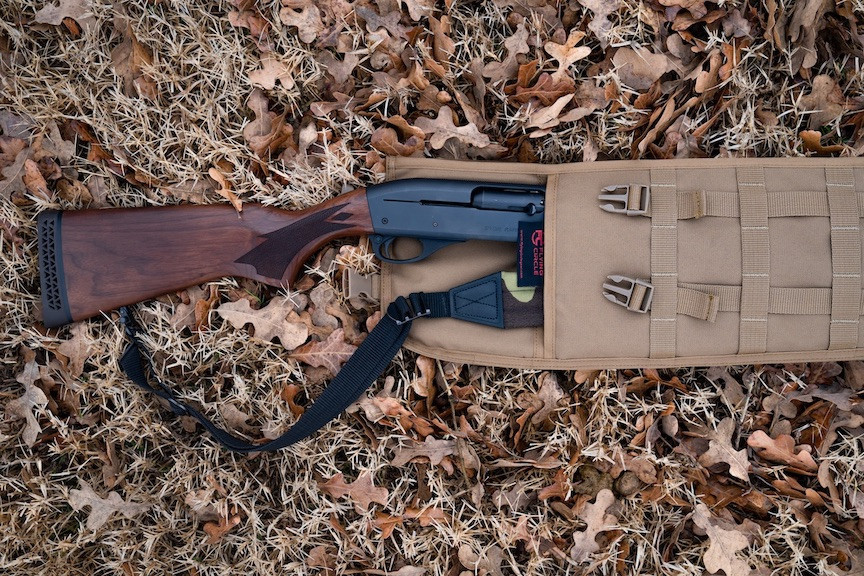 The top end of a Coyote Brown Shotgun Scabbard on the ground with brown leaves. There is a brown and black rifle inside the scabbard and the stock is seen outside of the top.