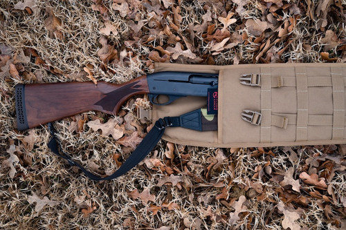 The top end of a coyote brown Shotgun Scabbard on the ground with brown leaves.  There is a brown and black rifle inside the scabbard and the stock is seen outside of the top.