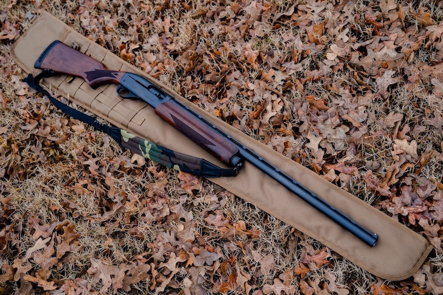 A Coyote Brown Shotgun Scabbard on the ground with brown leaves with a brown and black rifle with camo and black strap laid on top of it.