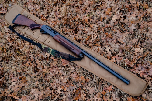 A Coyote Brown Shotgun Scabbard on the ground with brown leaves with a brown and black rifle with camo and black strap laid on top of it.