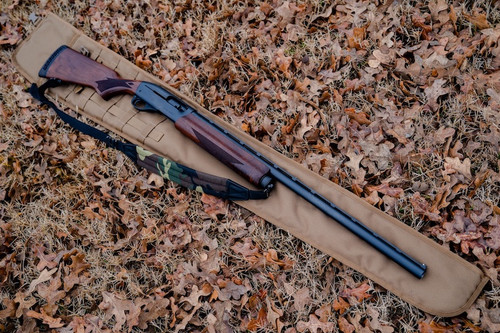 A coyote brown Shotgun Scabbard on the ground with brown leaves with a brown and black rifle with camo and black strap laid on top of it.
